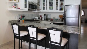 Una cocina moderna con armarios blancos, un refrigerador de acero inoxidable, un microondas y una encimera de granito. Hay tres taburetes de bar con asientos acolchados.