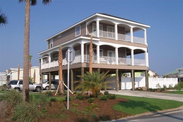 A two-story beachfront house with a balcony, surrounded by palm trees and a well-maintained lawn.
