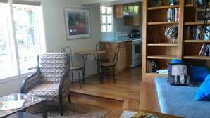 Een gezellige woonkamer met een bank, een salontafel en een gestreept fauteuil. Er is een keukenhoek met houten kastjes en een eettafel.