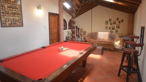 Ein Billardtisch in einem Raum mit hohen Decken und freiliegenden Balken, ein Korbstuhl und ein hölzerner Barhocker.