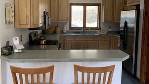 A kitchen with wooden cabinets, a stainless steel refrigerator, a large island with two chairs, and a window providing natural light. - Life is Good Lodge Montana on Gary Cooper Ranch at Dearborn River (Cascade)