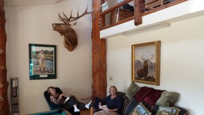 A cozy living room with a pool table, two people seated on a couch, and a mounted deer head on the wall. - Life is Good Lodge Montana on Gary Cooper Ranch at Dearborn River (Cascade)