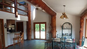 A spacious living area with a high ceiling, wooden beams, a dining table with chairs, a kitchen area, and a large window providing natural light. - Life is Good Lodge Montana on Gary Cooper Ranch at Dearborn River (Cascade)