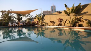 Una piscina con sillas de descanso y una vela de sombra, rodeada de plantas tropicales y un edificio al fondo.