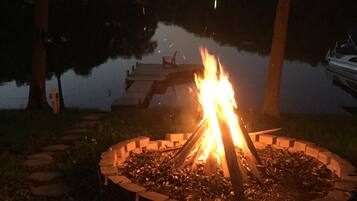 A campfire surrounded by a stone fire pit, with a dock extending into a calm lake.