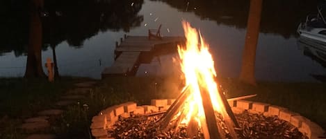 A campfire surrounded by a stone fire pit, with a dock extending into a calm lake.