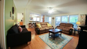 Un vaste salon comprenant un canapé en cuir marron, un fauteuil assorti et une table basse. On y trouve également un ventilateur de plafond, un tapis et une fenêtre avec des stores.