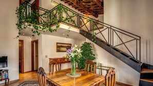 Un coin repas avec une table en bois, des chaises et un vase de fleurs. On y trouve un escalier avec une rampe en métal et une plante suspendue.