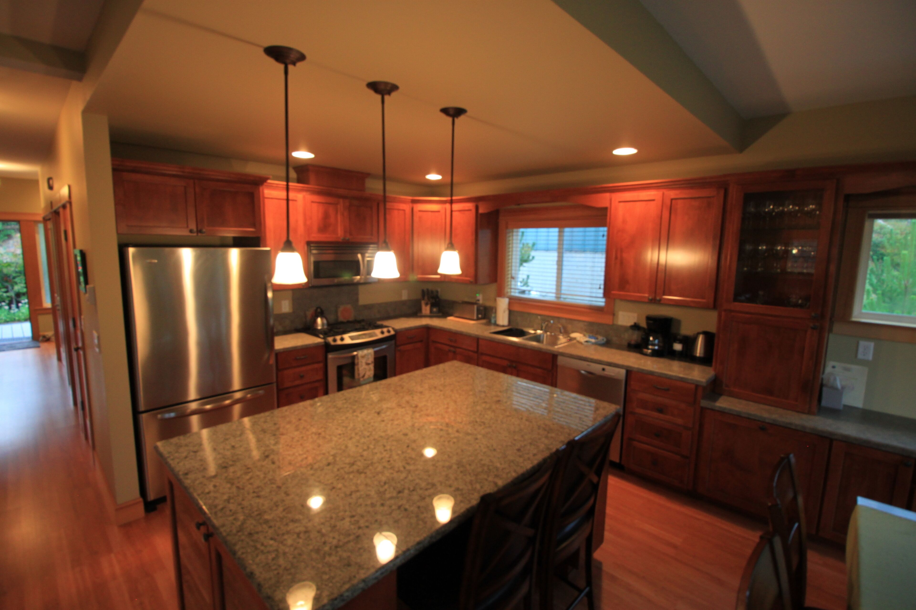 A modern kitchen with granite countertops, stainless steel appliances, wooden cabinets, pendant lighting, and a large island.
