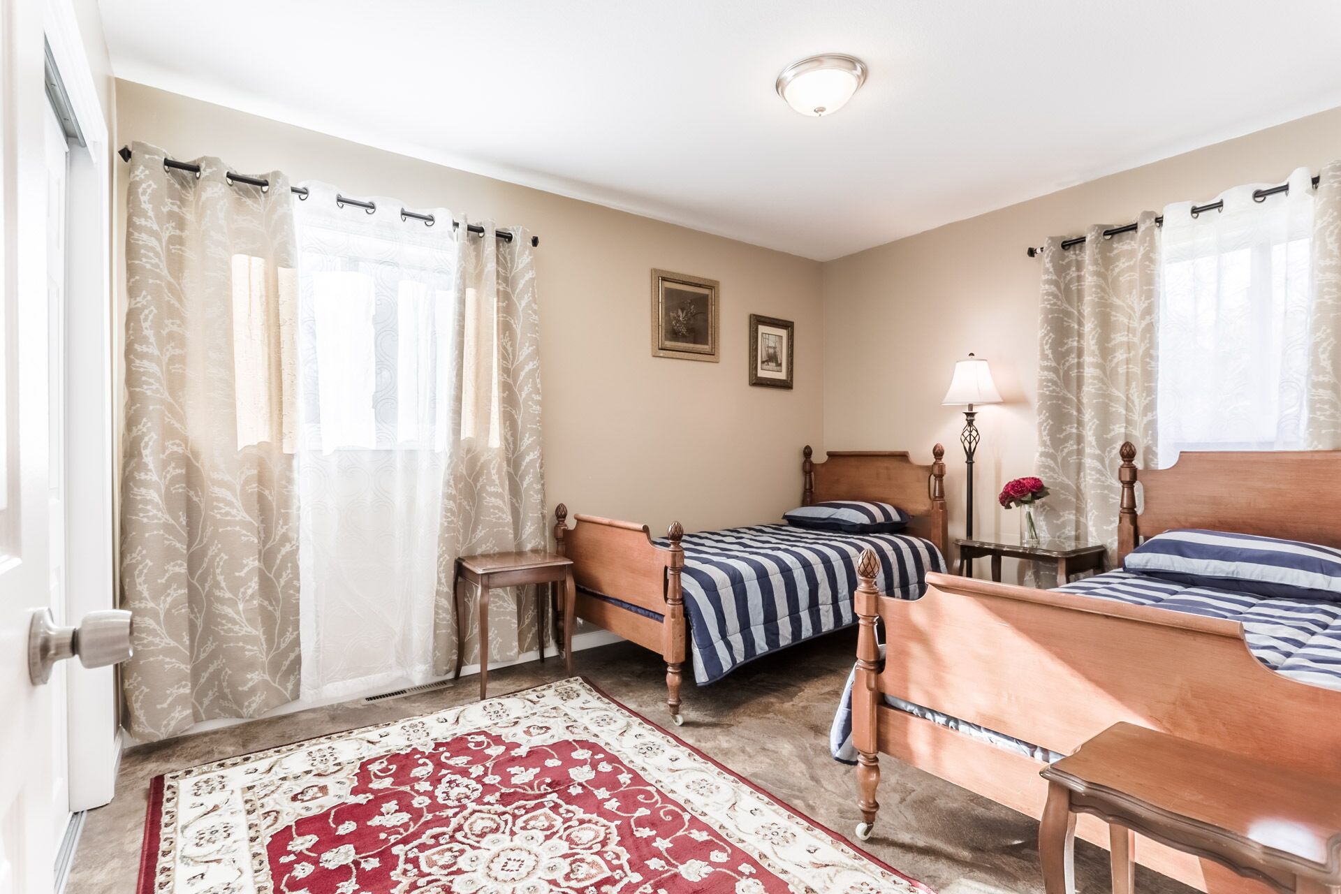 A hotel room with two beds, a wooden headboard, a bedside table with a lamp, a red and white patterned rug, and sheer curtains with a leaf pattern.