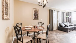A dining area with a round table, four chairs, and a chandelier. There is a sofa and curtains in the background.