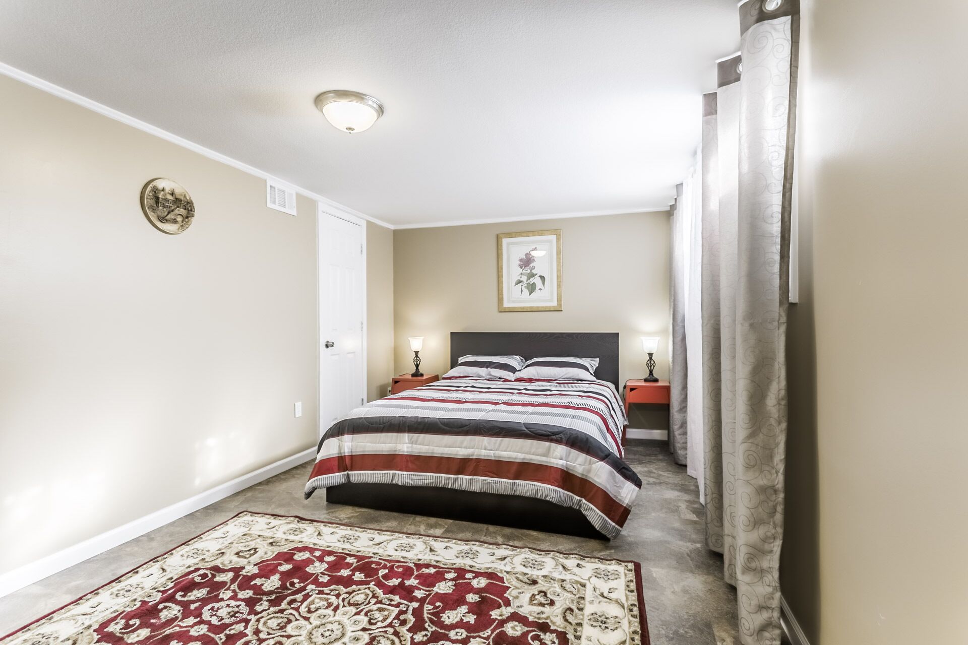 Ein Schlafzimmer mit einem Bett, einem Nachttisch, einer Lampe, einem gerahmten Bild an der Wand und einem gemusterten Teppich.