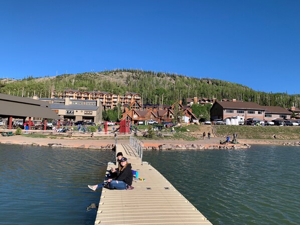 A pier extending into a lake with a backdrop of lodge buildings and a forested hill.