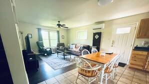 A living room with a sofa, chairs, a coffee table, and a TV. There is a dining table with chairs and a ceiling fan.