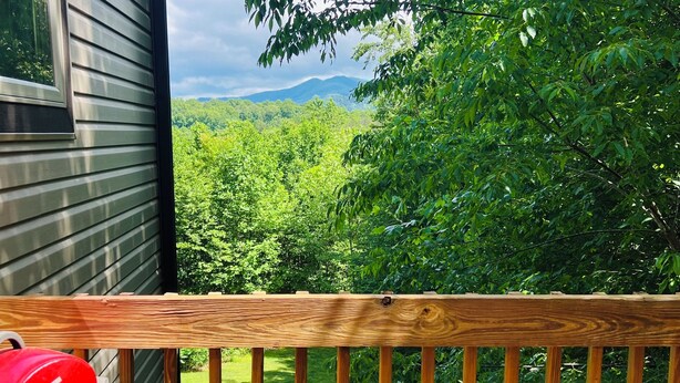 A wooden deck with a barbecue grill, overlooking a lush green landscape and a mountain in the distance.