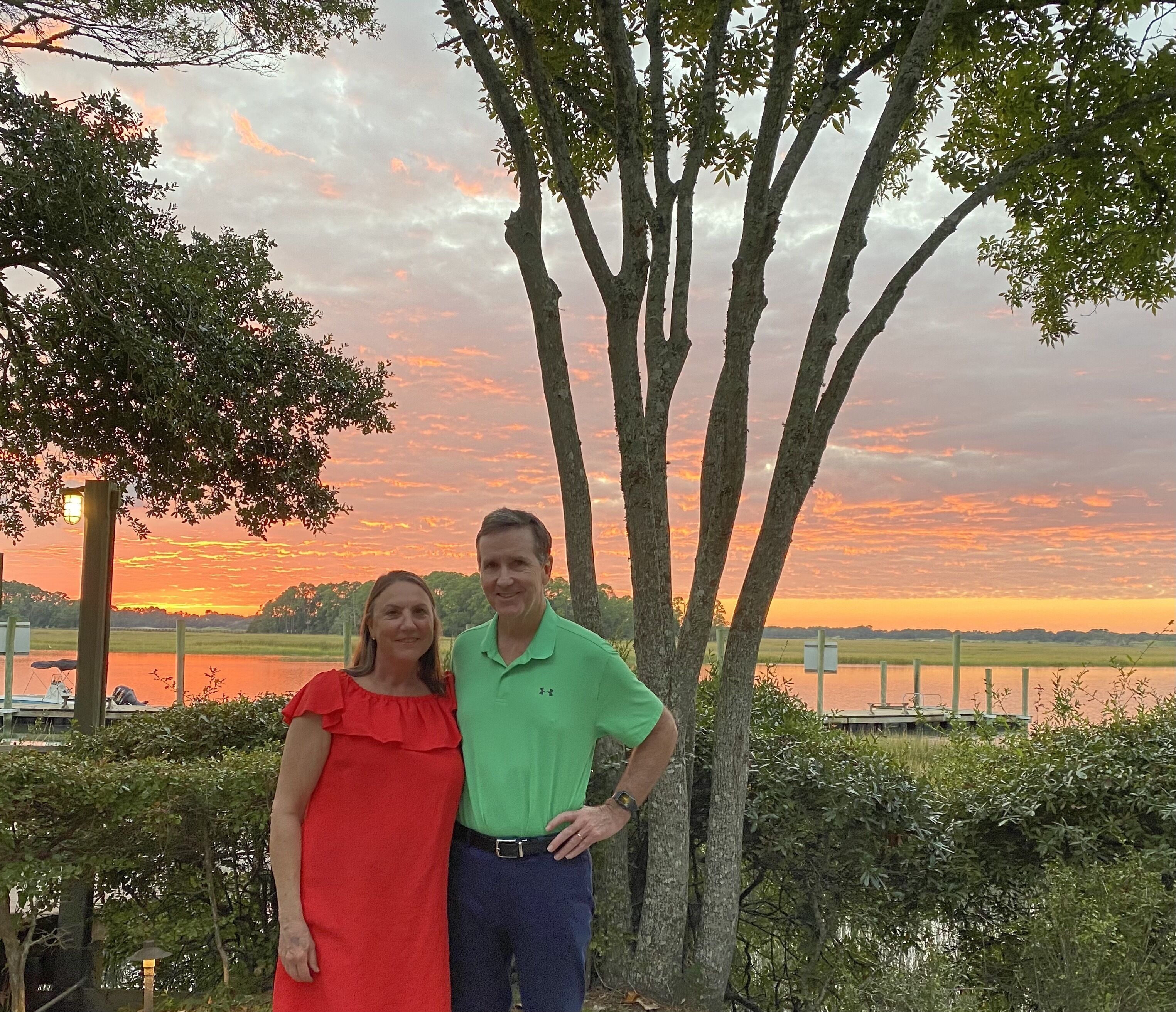 A couple standing by a lake with trees and a sunset in the background.