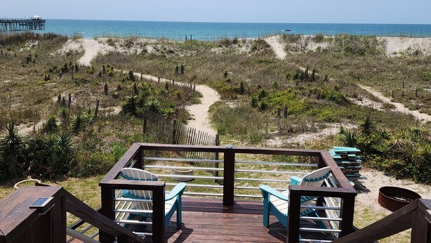 A beach view with a wooden deck, chairs, and a path leading to the ocean.
