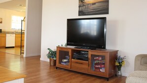 A living room with a flat-screen TV on a wooden cabinet, a painting of a sunset over water on the wall, and a kitchen in the background.