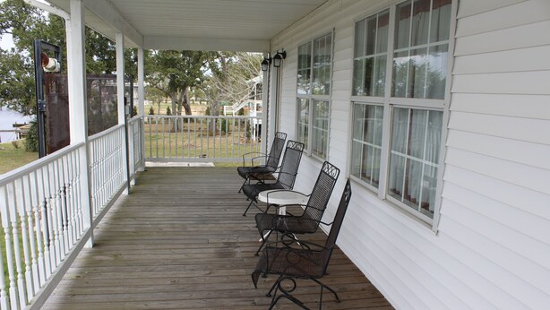 A covered porch with a wooden floor and white railings, featuring three metal chairs and a small table.
