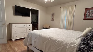 A bedroom with a bed, a dresser, a television, a ceiling fan, and a window with curtains.