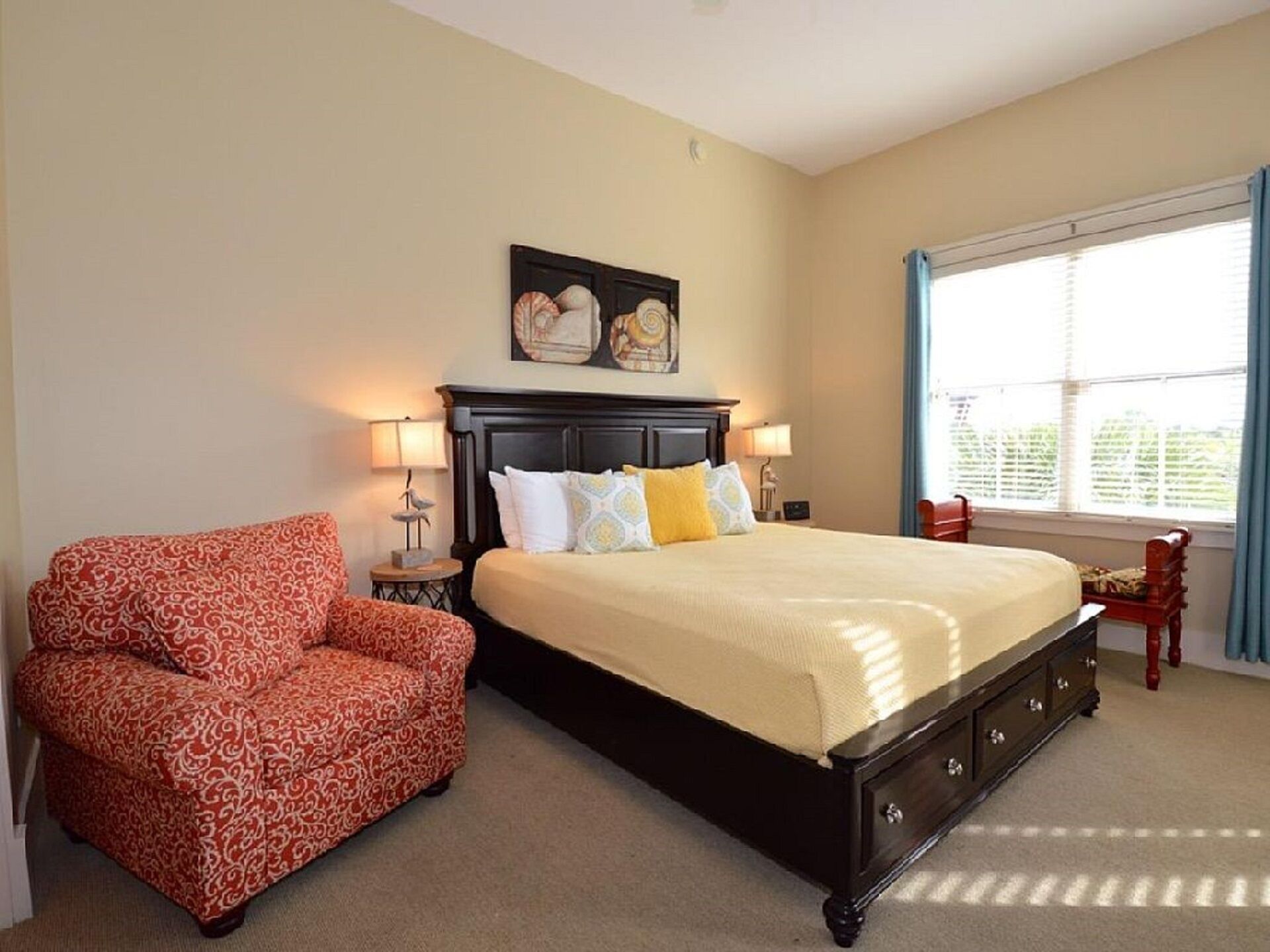 A bedroom with a large bed, two bedside lamps, a red patterned chair, a wooden nightstand, and a window with curtains.