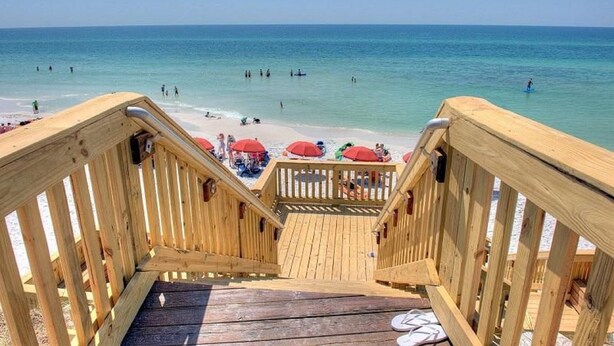 Eine Holztreppe führt zu einem Strand mit roten Sonnenschirmen und badenden Personen.