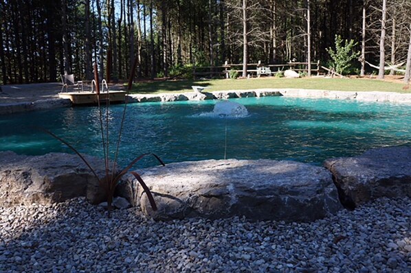 Una piscina con una fuente de agua, rodeada de rocas y guijarros, con un muelle de madera y una zona de césped al fondo.