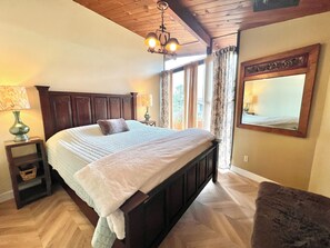 Quarto com uma cama grande, cabeceira de madeira, duas mesas de cabeceira com luminárias, um espelho e vista para o exterior por uma janela com cortinas.