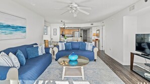 A modern living room with a blue sectional sofa, a glass coffee table, and a ceiling fan. - Oceanfront End Unit 4BR 3BA Sleeps 12 75 Inch TV Crescent Shores N Myrtle Beach (North Myrtle Beach)