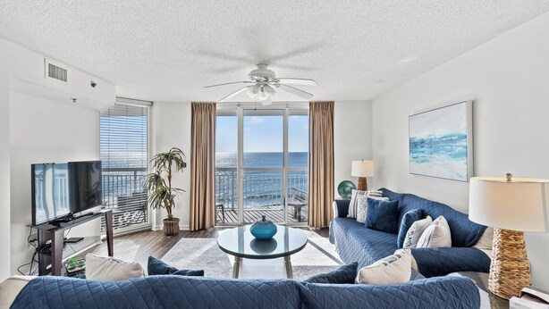 A modern living room with a large TV, a ceiling fan, a balcony with a view of the sea, and a cozy seating area with blue cushions. - Oceanfront End Unit 4BR 3BA Sleeps 12 75 Inch TV Crescent Shores N Myrtle Beach (North Myrtle Beach)