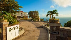 A coastal property with a paved driveway leading to a house with a red roof, surrounded by lush greenery and a view of the ocean.