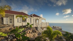 A coastal house with a stone foundation, white exterior, and a red roof. It features a balcony with a railing and a view of the ocean.