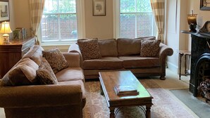 Un salon avec un ensemble de canapés marron, une table basse en bois et un tapis à motifs. On y trouve un ventilateur de plafond, une cheminée et une fenêtre avec des rideaux.