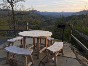 Un espace extérieur aménagé avec des meubles en bois, comprenant une table ronde et des bancs, le tout entouré d’une clôture en bois et offrant une vue sur un paysage montagneux.