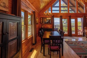 Interior de una cabaña de madera con una mesa de comedor, sillas, una araña y ventanas amplias que ofrecen vistas al exterior.