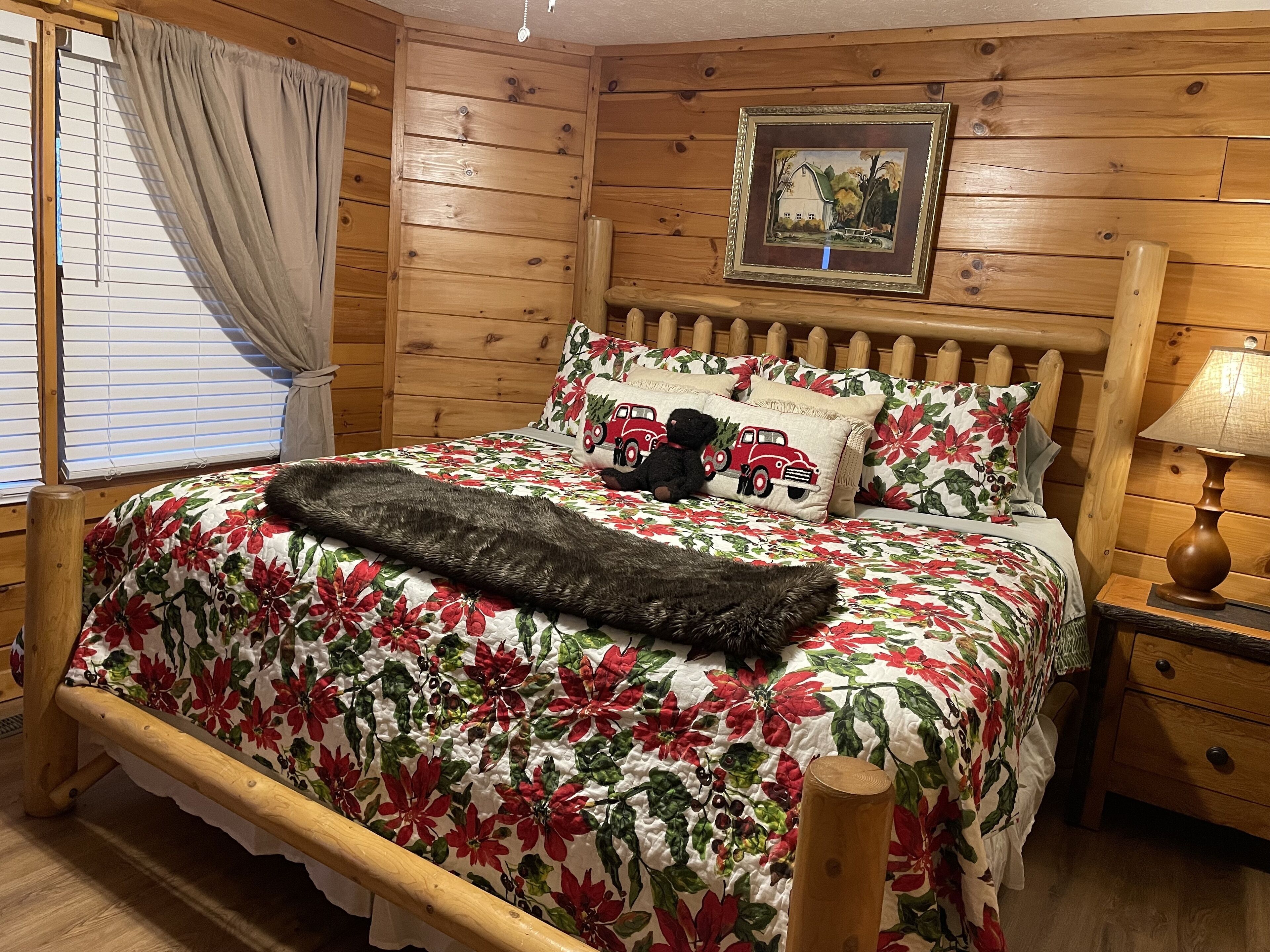 A wooden cabin-style bedroom with a bed, floral bedding, a teddy bear, a framed picture, and a nightstand with a lamp.