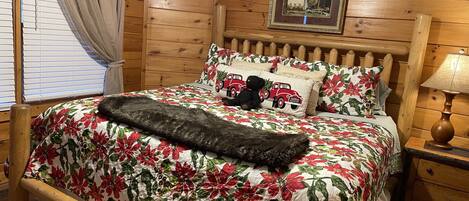 A wooden cabin-style bedroom with a bed, floral bedding, a teddy bear, a framed picture, and a nightstand with a lamp.