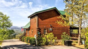 Une cabane en rondins surmontée d’un toit végétalisé, entourée d’une végétation luxuriante et avec une chaîne de montagnes en arrière-plan.