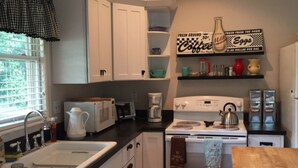 Una cocina con armarios blancos, un fregadero, una estufa y una cafetera. Hay una estantería con un letrero que dice “Coffee recién molido” y una botella de leche.