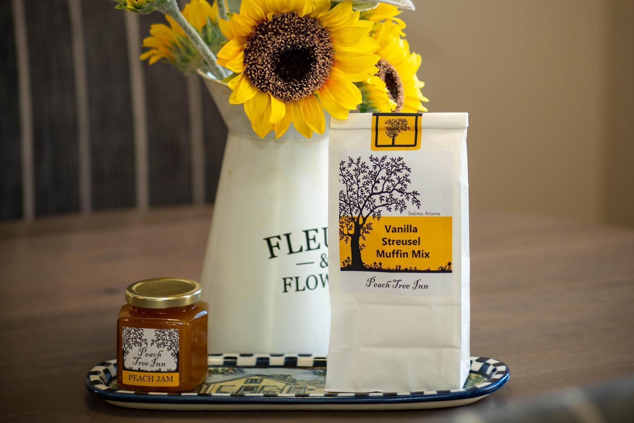 A jar of peach jam, a bag of vanilla streusel muffin mix, and a vase with sunflowers on a table.