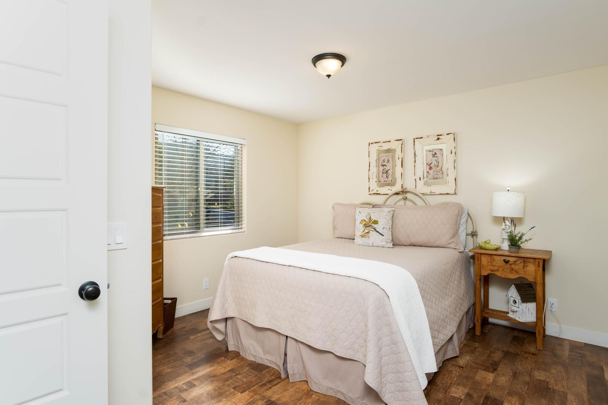 A bedroom with a bed, a nightstand, a window with blinds, and two wall-mounted pictures.