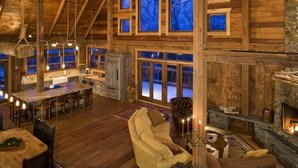 A spacious log cabin interior with a large dining area, a cozy living room with a fireplace, and multiple windows offering views of the outdoors. - Book for fall and winter now… dates are filling up!! (Maple)