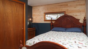 Une chambre à coucher avec une tête de lit en bois, une fenêtre donnant sur des arbres, une table de chevet en bois avec une lampe, et un lit recouvert d’une couvre-lit à motifs.