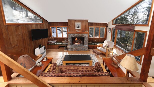 Un salon chaleureux avec une cheminée en pierre, des murs en bois et de grandes fenêtres donnant sur l’extérieur.