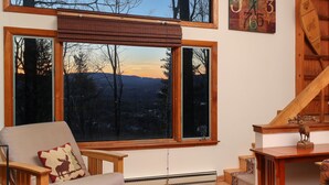 Une pièce dotée d’une grande fenêtre donnant sur un paysage montagneux, une chaise en bois avec un coussin orné d’un motif d’élan, une table en bois sur laquelle est posée une statuette de cerf, et un escalier en bois.