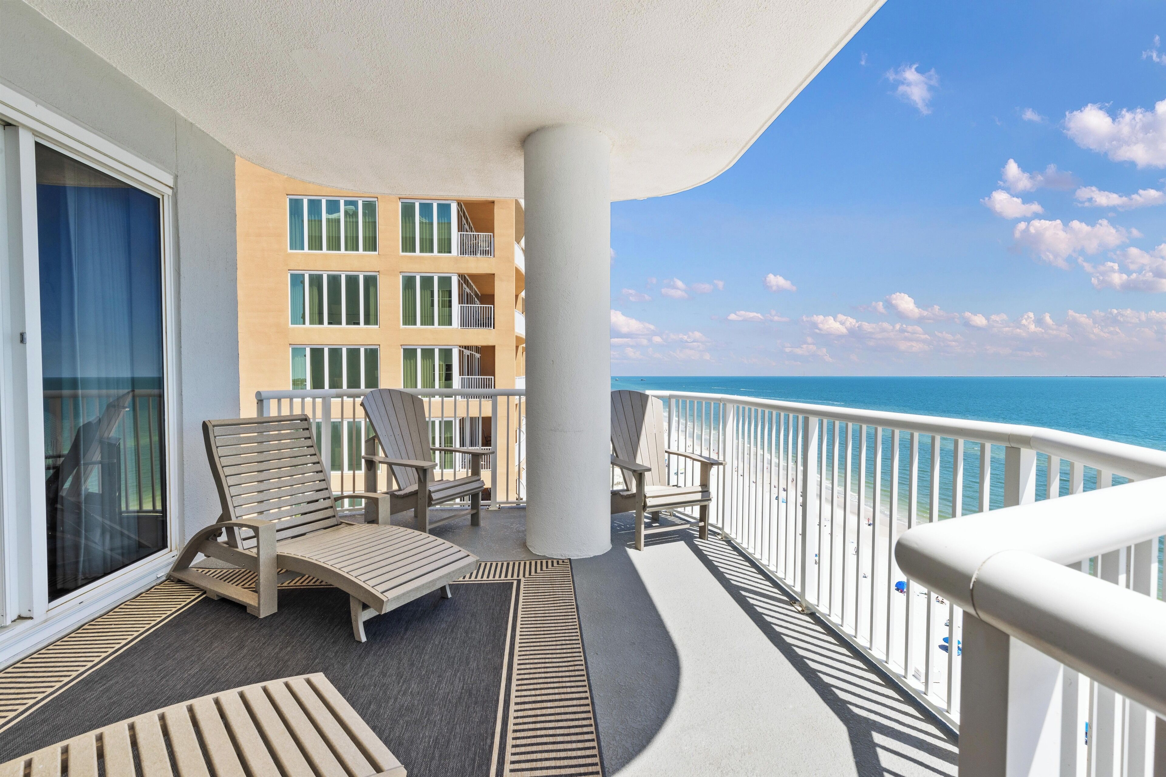 A balcony with a view of the ocean, furnished with chairs and a table, and a building with multiple balconies in the background.