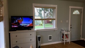 Una sala de estar con televisor, una ventana con persianas, una puerta blanca y una pequeña mesa blanca con libros.