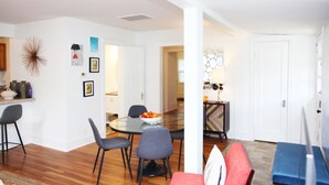 Una sala de estar moderna con una mesa de comedor de vidrio, sofás grises y rojos, y una zona de cocina con armarios blancos y salpicadero.