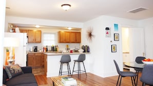 Una sala moderna con un sofá, una mesa de centro y un comedor con mesa y sillas. Se ve una cocina al fondo con armarios de madera y un ventanal.