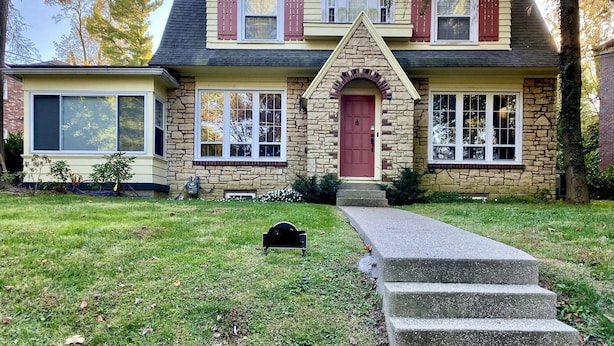 Una casa de dos pisos con fachada de piedra, una entrada central con puerta roja y un pequeño jardín frontal con buzón y escalones que conducen a la puerta.